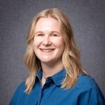 A smiling blonde woman wearing a blue button-up shirt against a gray background.