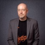 A man with glasses wearing blazer and graphic tee against gray background