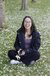 Photograph: woman sits cross-legged on grass with white petals, wearing black coat and purple top.