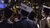 Three graduates wearing mortarboards and gowns facing a crowded graduation ceremony audience