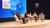 Five panelists seated on a round stage with a blue world map projection behind them