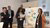 Five presenters in business attire discuss a flipchart with colored thought bubbles at a meeting