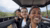 Three friends in an open-top safari jeep wearing jackets, green shrubs behind