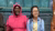 Two women on a bench before a teal shutter. One in pink tracksuit holds a water bottle; the other wears denim.
