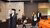 Academic official in gown with ceremonial staff leading three people past orange auditorium seats