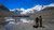 Two hikers stand at a rocky glacial lake, snow-capped mountains mirrored in calm water