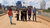 Six casually dressed people walk along a dusty urban road beneath a cloudy sky.