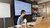 Photograph: woman at meeting table reviewing laptop with projected slides behind