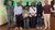 Nine casually dressed individuals standing and smiling in front of green-lit wall indoors