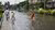 People wading through knee-deep floodwater on city road past apartment buildings