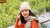 Young woman wearing a white beanie and plaid vest, sitting at a wooden table by a flower field.