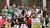 About 30 people smiling outdoors by a wooden pavilion, holding a green event sign