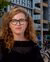 Woman with curly hair and glasses smiling in front of modern building and parked bicycles.